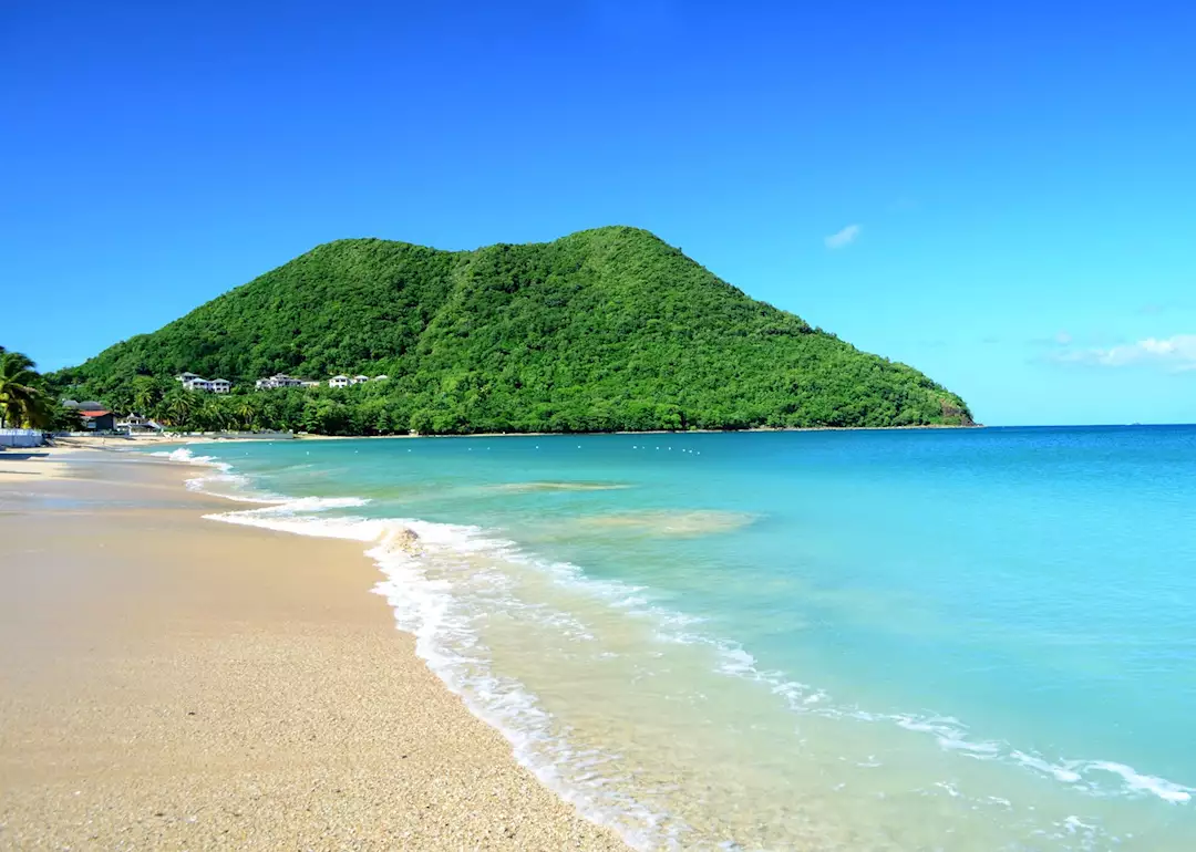 White sandy beach with waves washing ashore with clear blue waters and a lush green mountain coming up out of the water