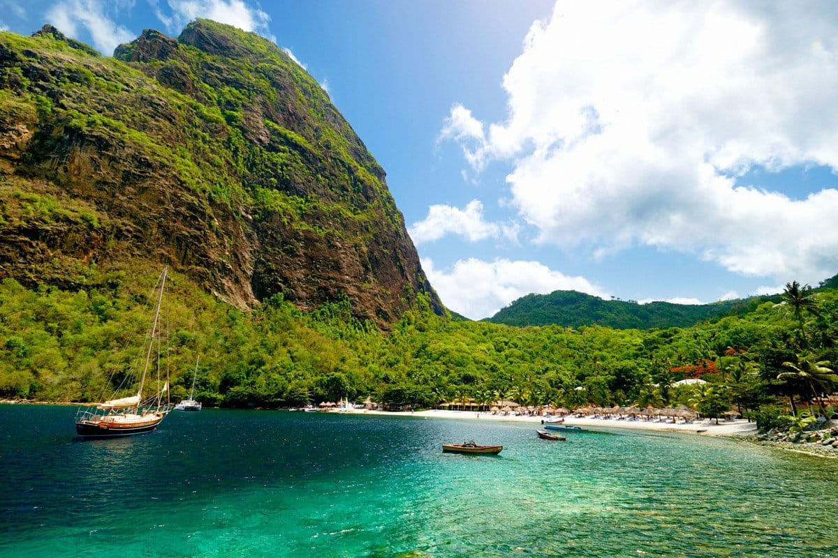 Beautiful beach with clear waters with sailboats anchored in the waters in front of the beach and a giant lush mountain next to the beach adding to it's beauty. 