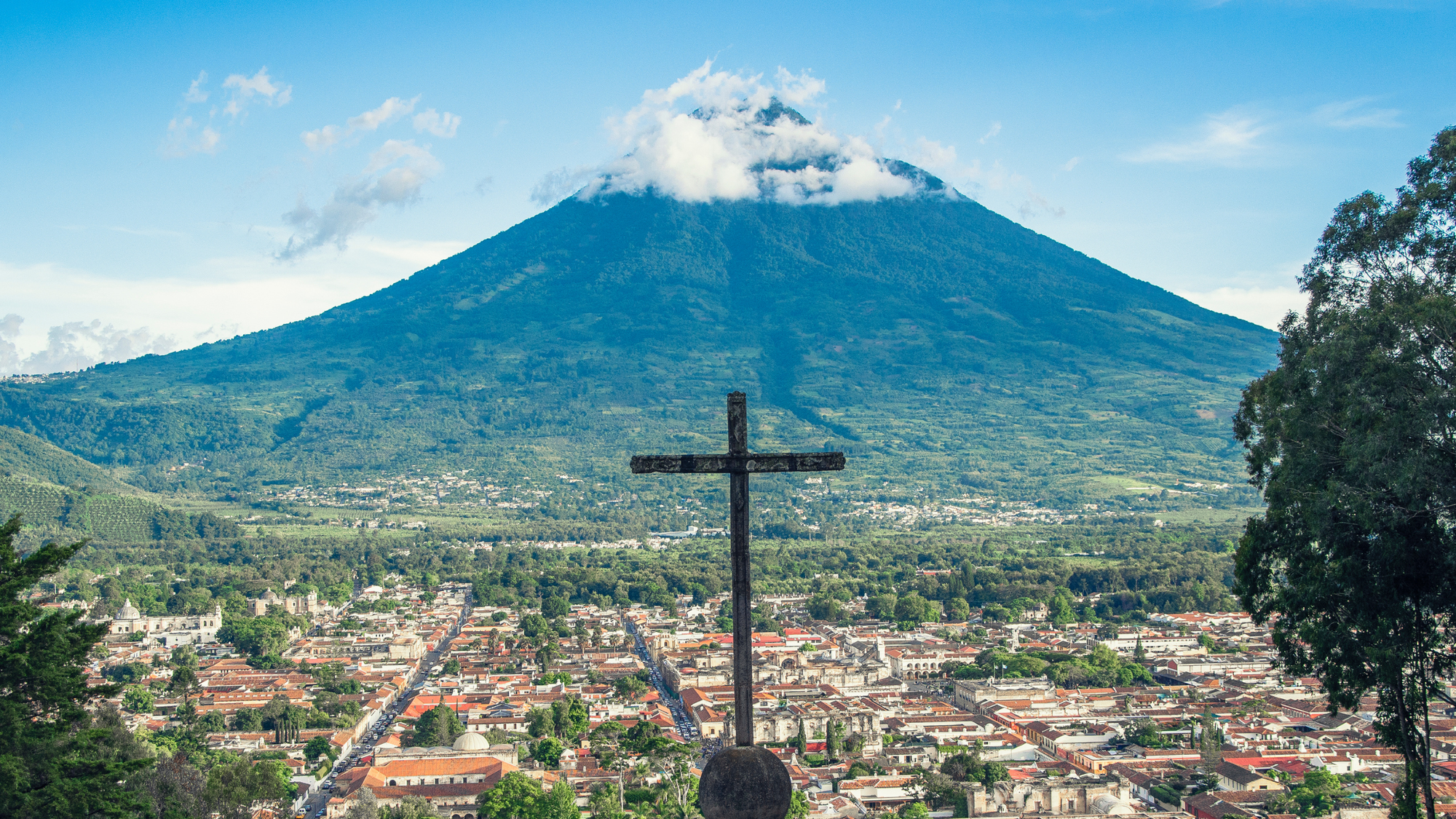 View of a cross seen on the hillside with a town sitting below and a large volcano in the background in Antigua, Guatemala