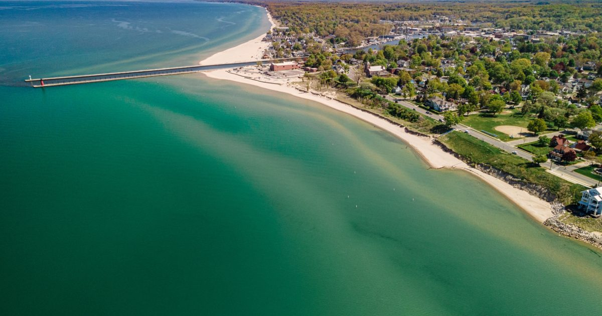 Ariel view of a white sandy beach with clear green waters with a pier leading out into the water at North Beach, South Haven Michigan.