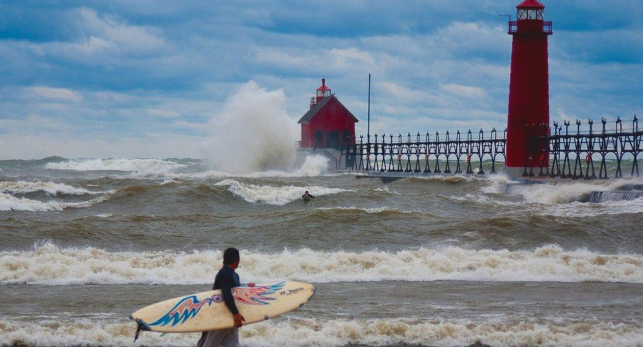 Pier with large waves and someone paddling on the board trying to catch a wave with another surfer holding their surf board walking on the beach watching them. 