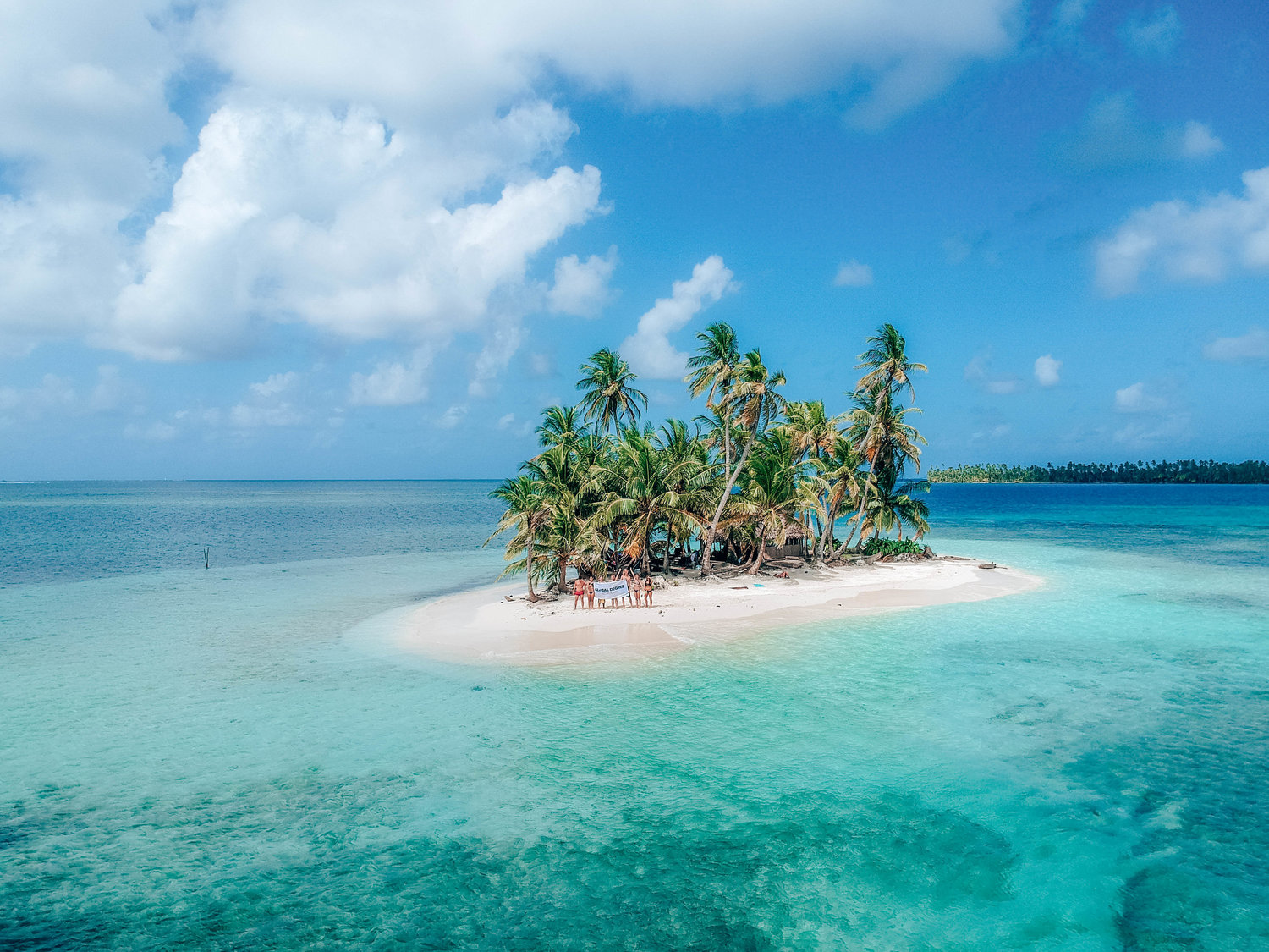 Little island with palm trees surrounded by clear waters and white sandy beach with people visiting the island.