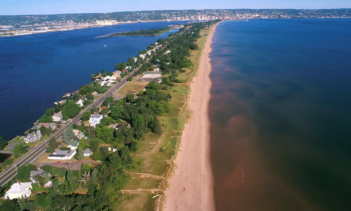 Park Point Beach on Lake Superior Ariel View of the beach that is a long beach that is lined with trees and water on both side of the beach and a road between the two bodies of water. 