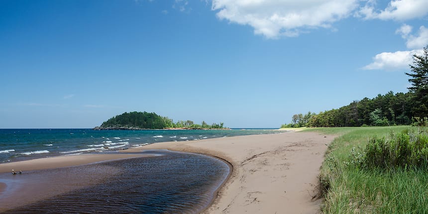 A tan sandy beach with small waves washing a on the shore line at Little Presque Isle Beach on Lake Superior.