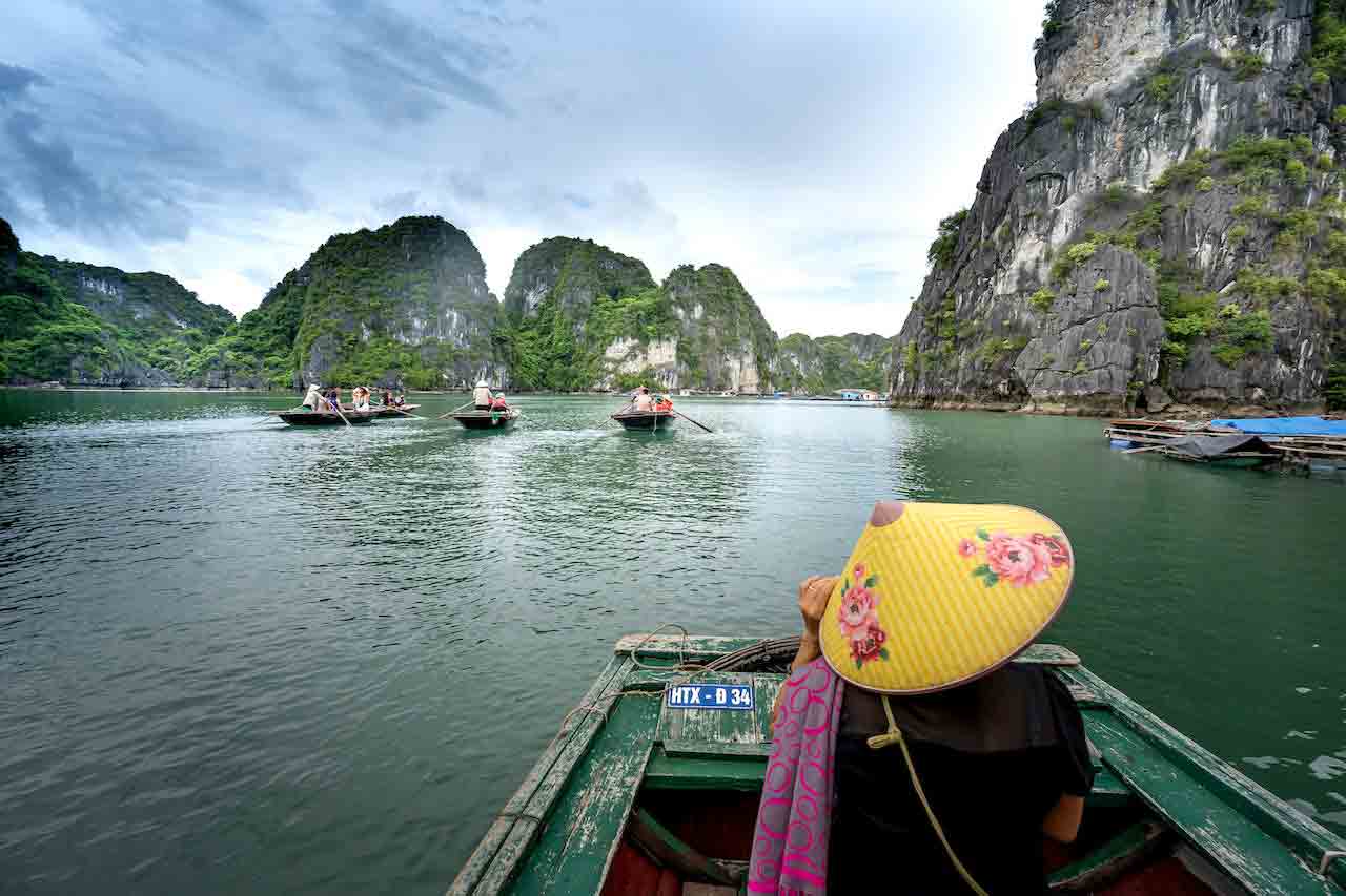 Native Vietnamese woman riding in front of a wooden boat surrounded by beautiful mountains that are covered in lush forest.