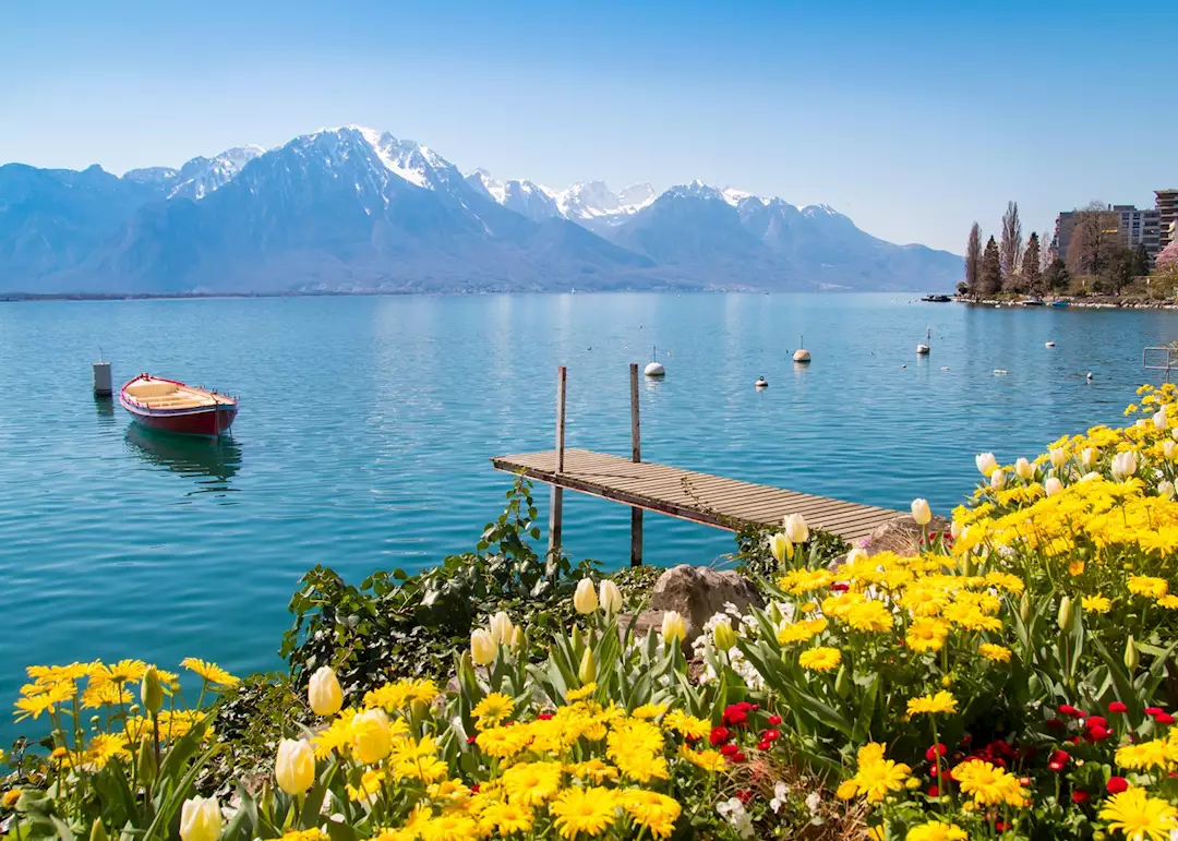 A scenic view of Lake Geneva, Switzerland, featuring a wooden pier, a floating boat, colorful flowers, and snow-capped mountains.