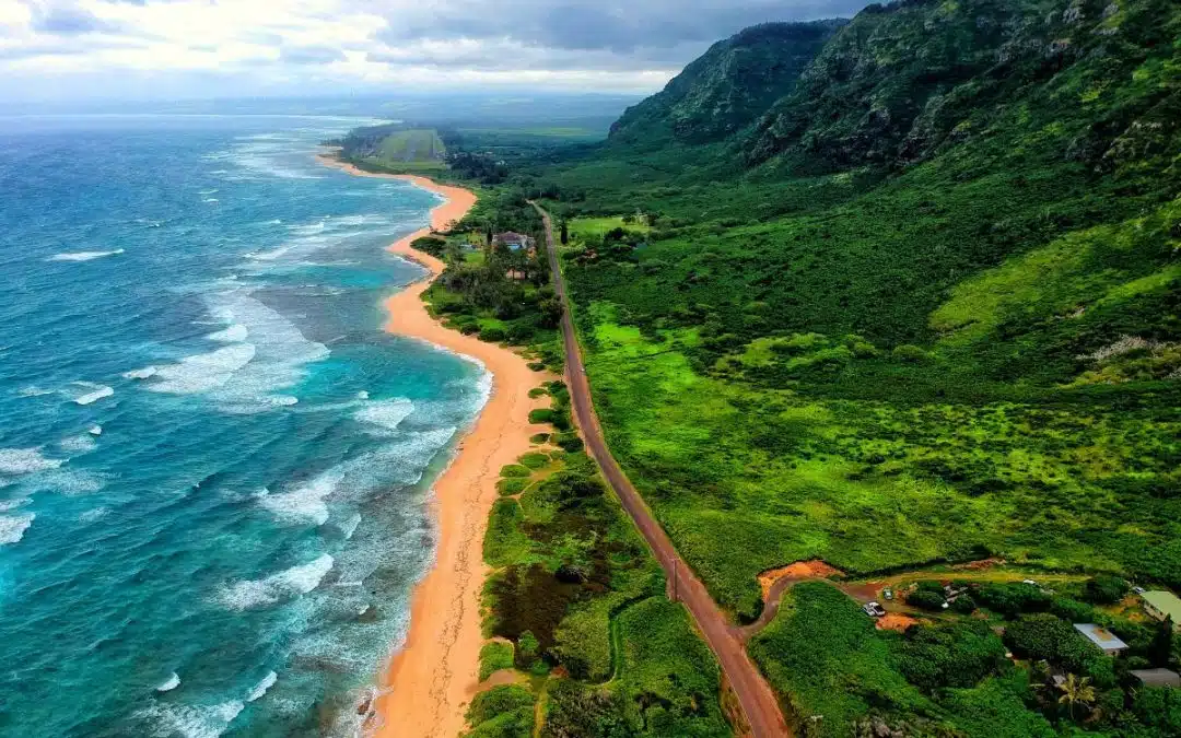 Ariel view of a beach with blue waters and waves breaking, and the beach has a road running behind it with large mountains covered lush green plants beyond the road as far as you can see. 