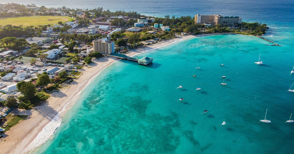 Ariel View of a beach with very clear blue waters and the beach has a few resorts that run along it, and the bay has sailboats anchored in the pretty waters. 