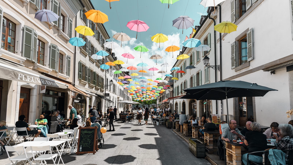 Bright colored umbrellas that have been placed in a way that appears they are floating above a busy street of people eating in the area of Carouge, Switzerland