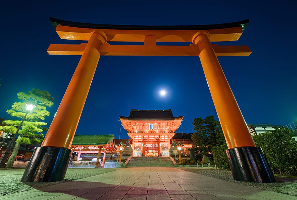 Entrance to Fushimi Inari Shrine is a giant red gate with Japanese styles to it with the building that holds the shrine in the distance that is lit up with red lights at night time.