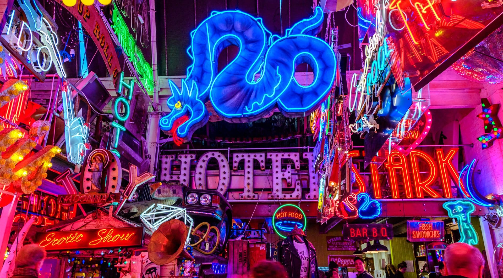 A vibrant and colorful scene from God’s Own Junkyard in London, UK, showcasing an eclectic collection of neon signs in various shapes, colors, and designs. A large blue neon dragon dominates the center, surrounded by glowing words like "HOTEL," "STARK," and "Erotic Show." The space is densely packed with vintage signs, illuminated artwork, and nostalgic memorabilia, creating a visually striking and retro atmosphere