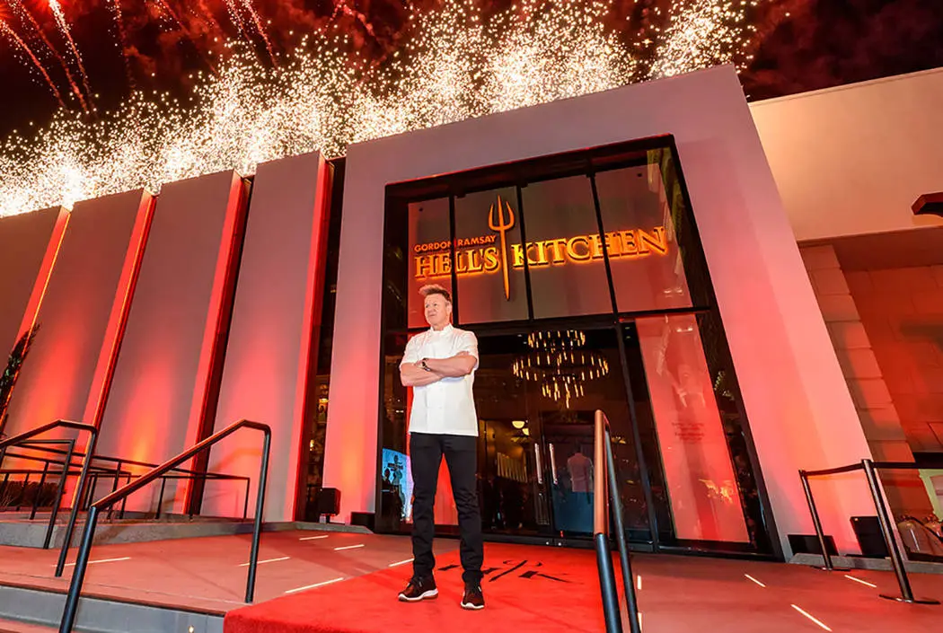 Gordon Ramsey standing outside of his Gordon Ramsay Hell’s Kitchen In Las Vegas with the front showing the sign saying Hells Kitchen and a devils 3 prong weapon on the sign lit up in red. 