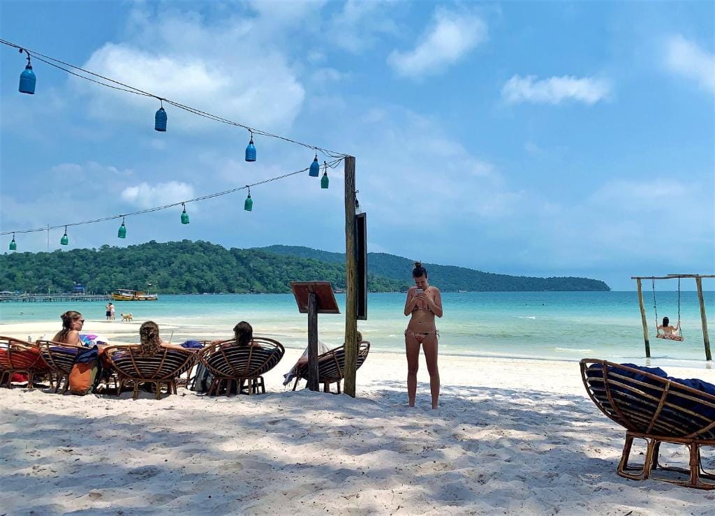 Women sitting in weaved chairs on a sandy beach with clear blue waters and a person swinging on a wooden swing that has been built at the waters edge enjoying themselves. 