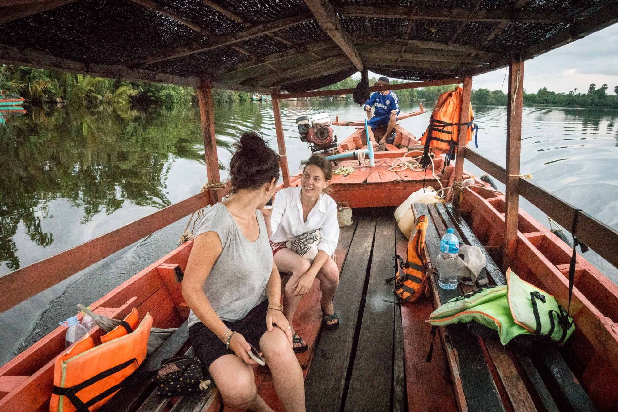 Two women riding on a wooden boat that is enclosed and has a local Cambodian man driving them around and giving them a tour of the river in Kampot.