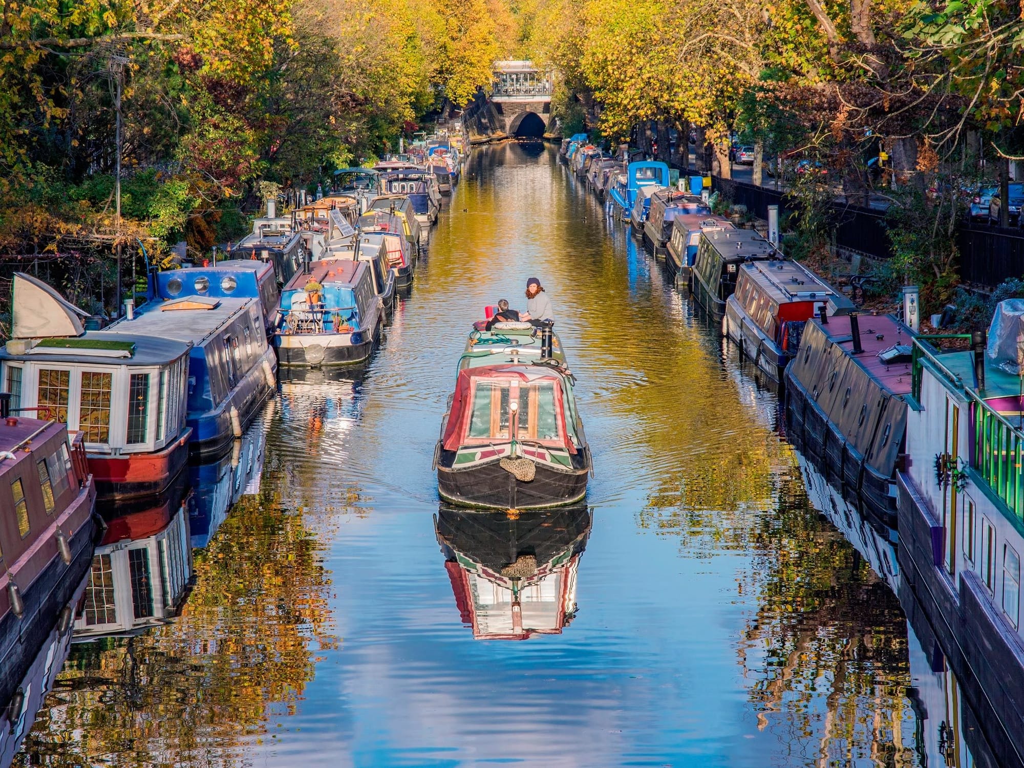 Narrow long house boat going up a canal with other similar boats parked all along the canals edge on both sides.