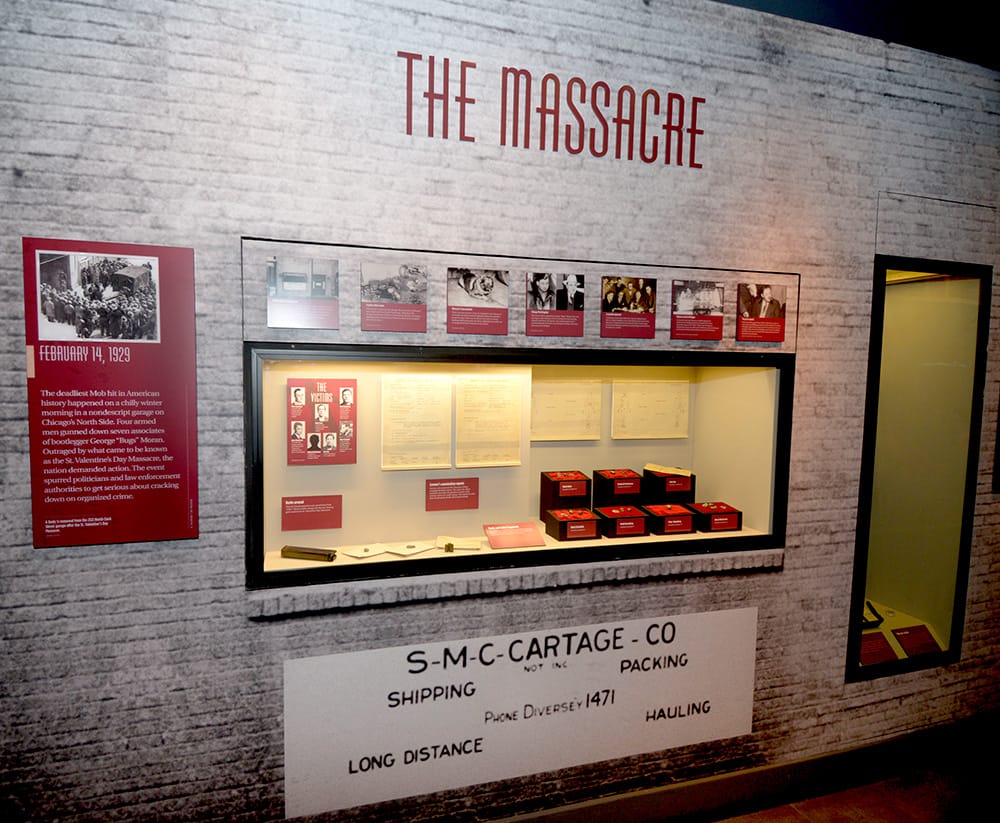 The Massacre display inside the mob museum in Las Vegas with a white brick wall with a glass display case displaying things pertaining to the mob killings. 