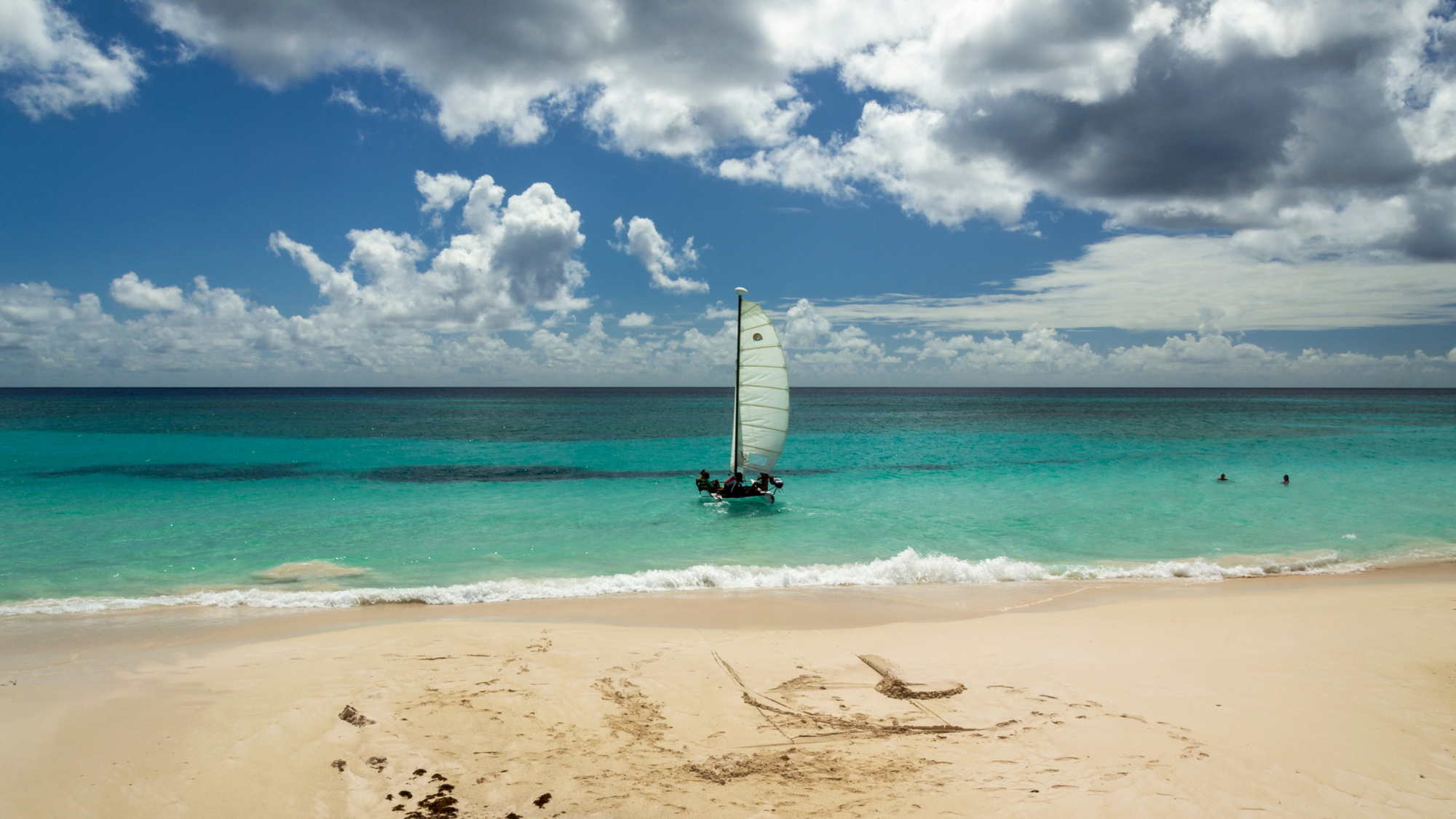 Lone small personal sailboat in clear Caribbean waters with a couple people on the boat just off a clean sand beach. 