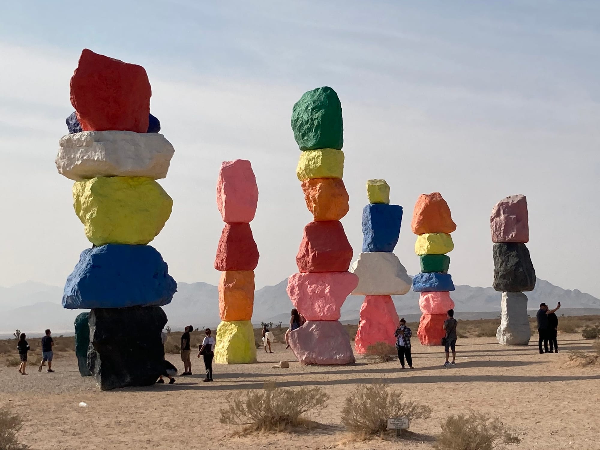 Colorful rocks of all different colors stacked on top of each other in the middle of the desert with people there taking photos. 