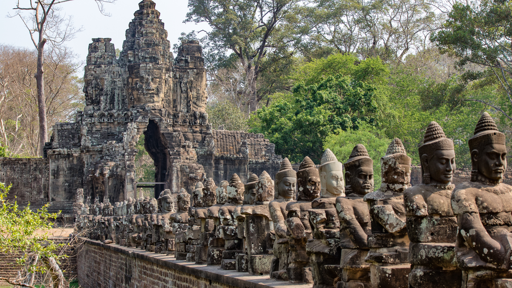 Old statues of buddha at at Angor Wat lined up on the side of a rock wall and a temple in the background. 