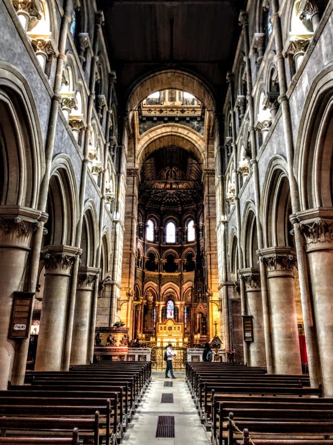 Interior of St Fin Barre’s Cathedral with benches for worshipers to sit at and someone is up front looking at the amazing work the builders did creating this cathedral. 