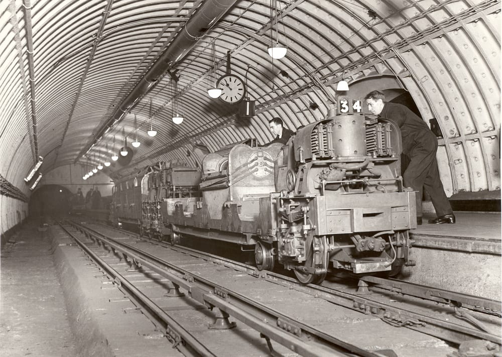 Black and white photo of two men working on an old train that is inside of an underground tunnel.