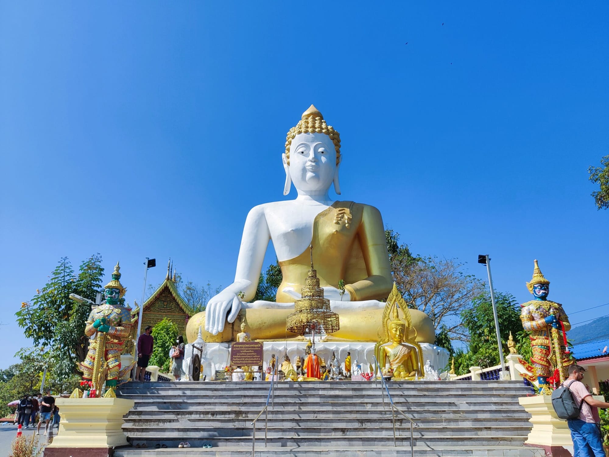 Giant White and Gold Buddha statue sitting with his legs crossed on top of stairs with tourist walking by. 