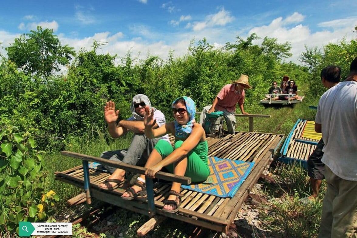 A man and a woman wearing sun protecting scarfs on their head while riding a bamboo train cart being operated by a Cambodian man, with another cart also giving tourist a ride behind it. 