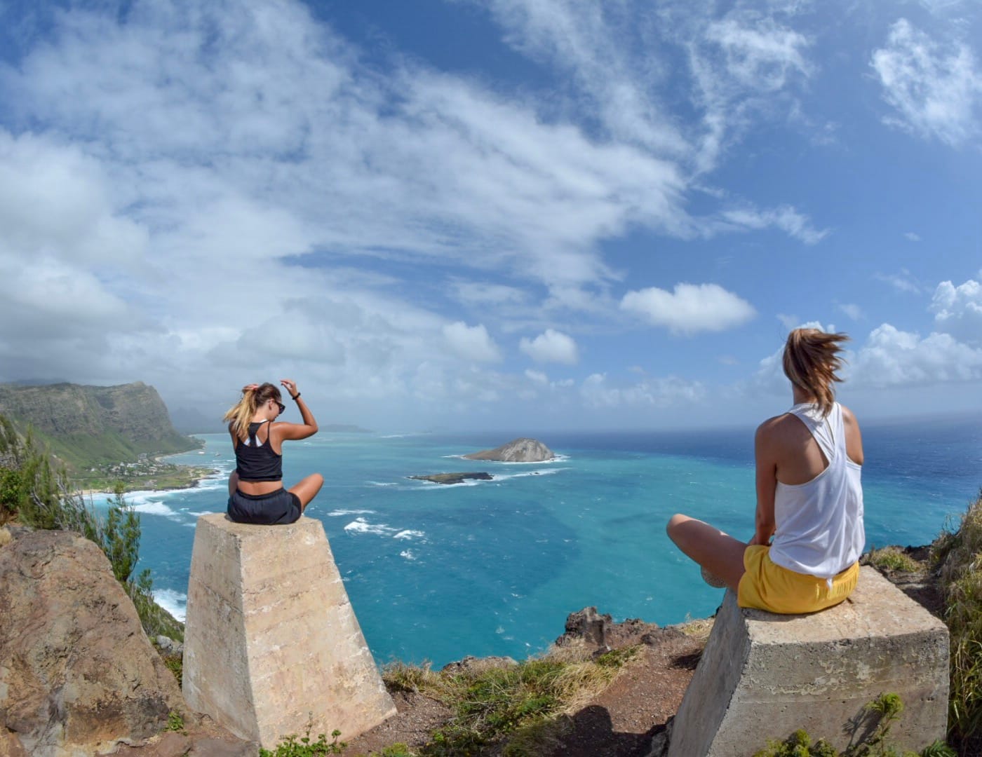 Two people sitting on top of concrete blocks that are at the top of a mountain over looking the ocean.