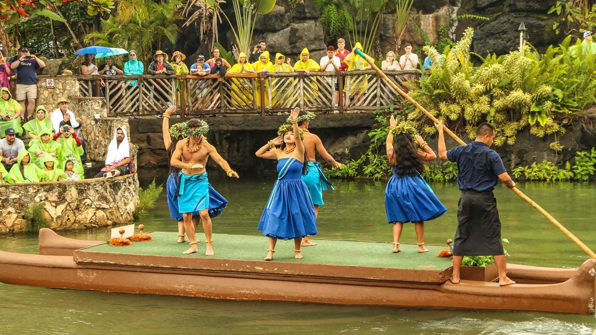 Native Hawaiian dancers dancing on a canoe being poled by another Hawaiian while spectators watch from a bridge. 