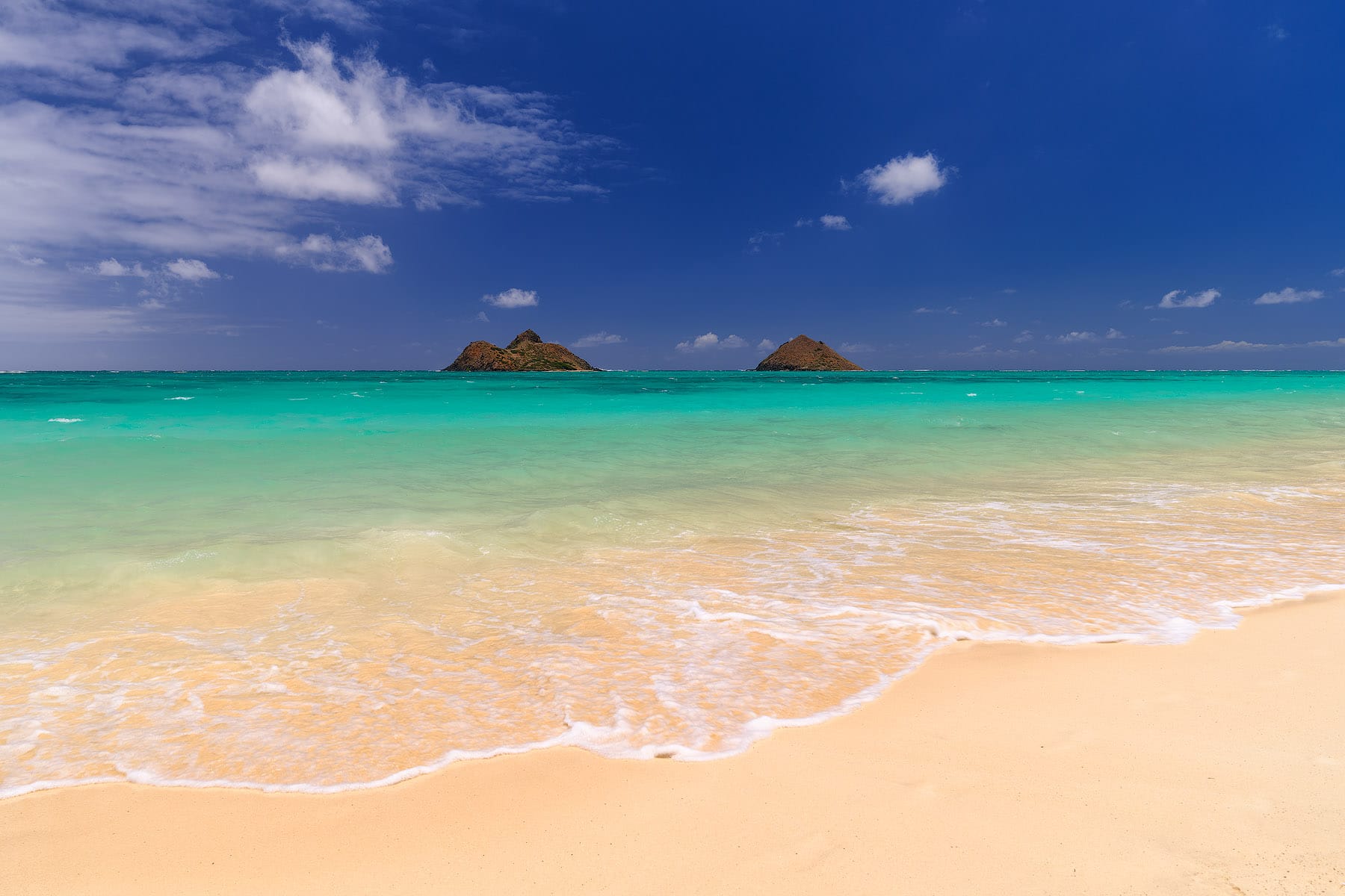 Two small islands out in the water off of a pretty beach with clear blue waters. 