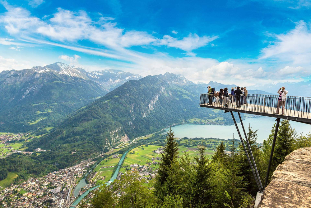 Tourists on a scenic lookout platform in Interlaken, Switzerland, overlooking mountains, a turquoise river, and a lake.