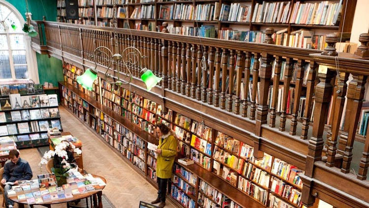 Man reading a book on the bottom level of a two level room where the walls are lined with books from floor to ceiling. 