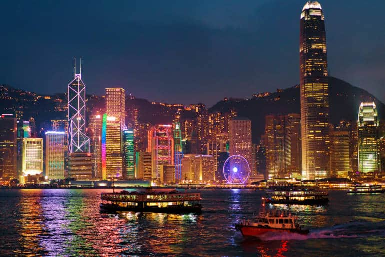 View of bright colored sky scrapers at night along the waterfront with ferry boats crossing across the water. 