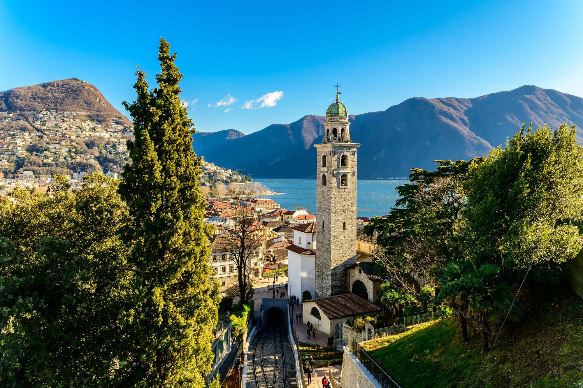 Scenic view of Lake Lugano, Switzerland, featuring a historic church tower, lush greenery, and mountains under a clear blue sky.