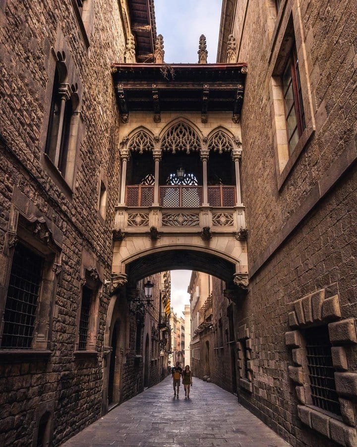 Pont del Bisbe (Bishop's Bridge) in Barcelona, Spain, a neo-Gothic stone bridge with intricate tracery and ornate details, arching over a narrow cobblestone street in the Gothic Quarter.