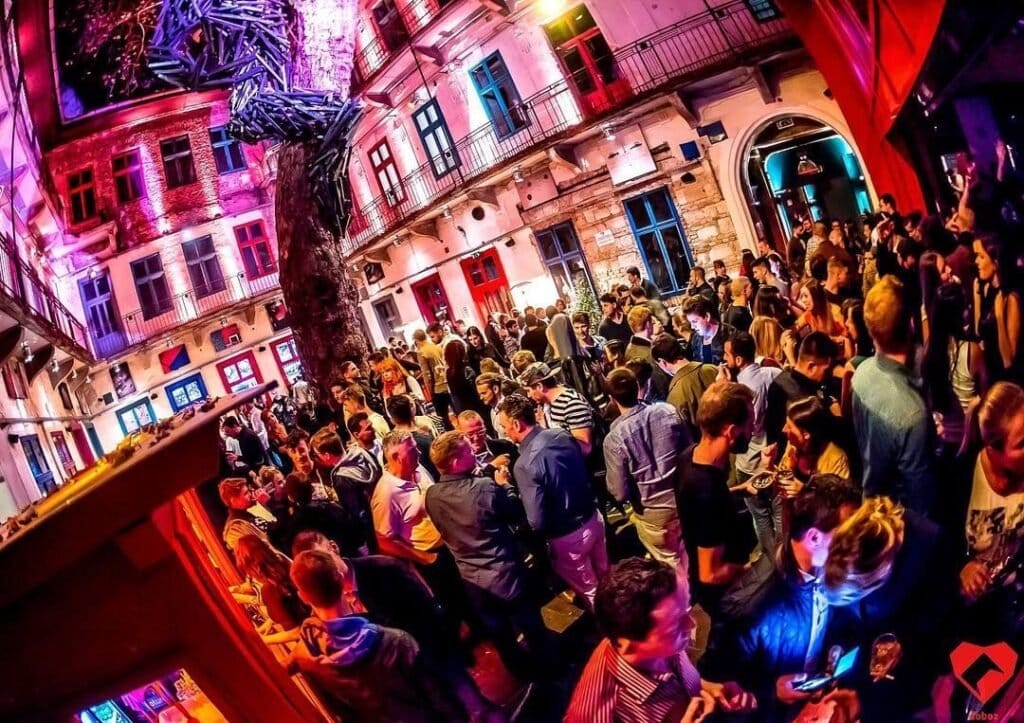 A nightclub in Budapest with a tree in the center of the courtyard with tons of people drinking and socializing. 